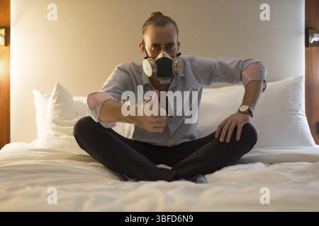 Hübscher junger, stilvoller kaukasischer Mann in Atemschutzmaske, der auf dem Bett im Schlafzimmer sitzt Stockfoto