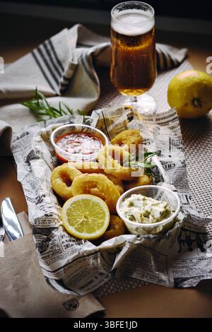 Nahaufnahme knuspriger Calamari-Ringe, serviert mit eintauchenden Soßen, Zitronenscheibe und Kräutern auf Zeitungspapier, mit einem Glas Bier im Hintergrund Stockfoto