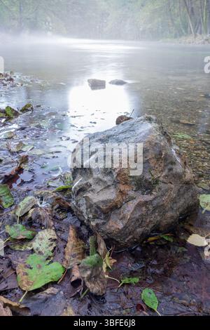 Quelle des Flusses Bosna, Sarajevo-Ilidza. Der Morgennebel schwingt über dem ruhigen Flusswasser, enthüllt einen großen Felsen und schafft eine ruhige Atmosphäre Stockfoto