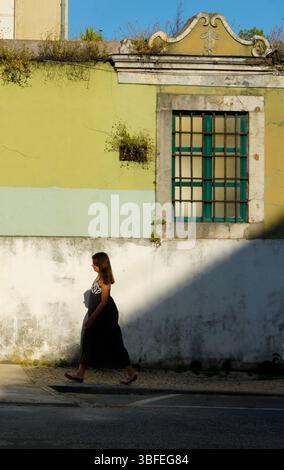 Frau geht an verwitterten gelben und weißen Wänden vorbei, mit grünen Fensterläden, die dramatische Schatten auf der kopfsteingepflasterten Straße im alten Lissabon werfen Stockfoto
