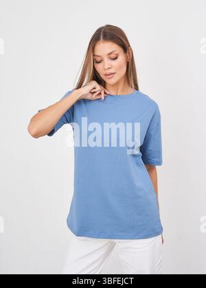 Junge Frau in einem schlichten blauen T-Shirt mit kurzen Ärmeln und weißer Hose posiert mit geschlossenen Augen und einem leichten Lächeln. Sie berührt ihren Hals mit einem Stockfoto