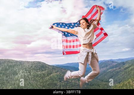 Eine junge Frau springt überschwänglich in die Luft und drückt ihren patriotischen Geist aus, während sie die amerikanische Flagge schwenkt. In lässiger weißer Kleidung steht sie Stockfoto