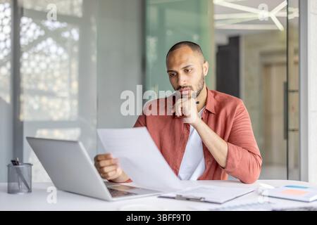 Ein fokussierter Mann liest Dokumente an seinem Schreibtisch in einer modernen Büroumgebung und berücksichtigt sorgfältig Details. Stockfoto