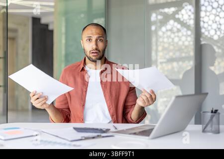 Ein überraschter Mann hält Papierkram, während er an seinem Schreibtisch arbeitet, und hat möglicherweise finanziellen Stress. Stockfoto