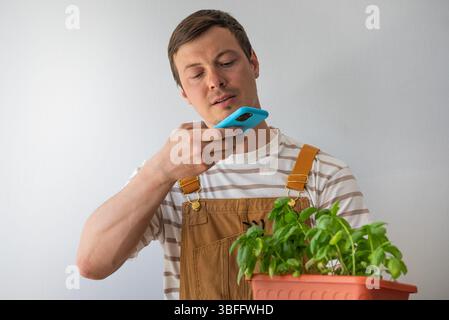 Junger Mann in Overalls, hält einen Topf frisches Basilikum in der Hand, während er auf seinem Smartphone eine Sprachnotiz aufnimmt, die die Freuden der Gartenarbeit und der Pflege von Pflanzen im Haus veranschaulicht Stockfoto