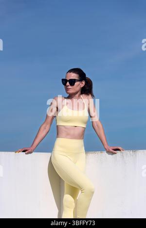 Stilvolle Frau mit gelber Aktivkleidung posiert selbstbewusst vor einer weißen Wand Stockfoto