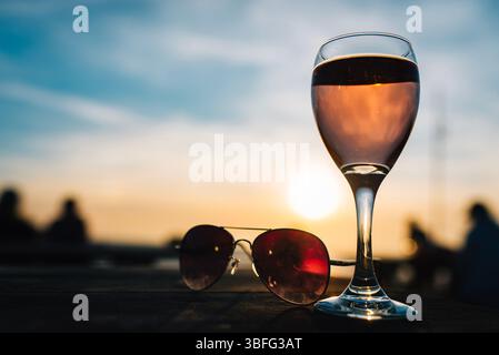 Ein Glas Rosé und eine Sonnenbrille auf einem Tisch am Strand bei Sonnenuntergang sorgen für eine entspannte Sommerabendstimmung mit warmen Tönen und einem weichen Hintergrund. Stockfoto