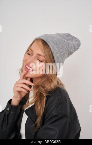 Fröhliche Frau mit grauer Mütze und Lederjacke posiert drinnen mit einem verspielten Lächeln Stockfoto