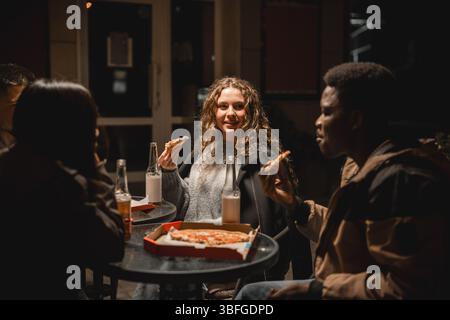 Eine Gruppe von Freunden genießt abends im Freien Pizza und Getränke in gemütlicher urbaner Umgebung Stockfoto