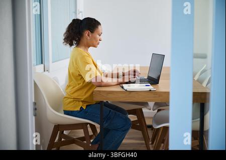 Konzentrierte Frau saß an einem Schreibtisch und arbeitete an ihrem Laptop in einem hellen, sauberen Home Office. Demonstriert Remote-Arbeiten, freiberufliches Arbeiten und Home-Office-Setups für Stockfoto
