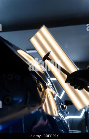 Der Techniker verwendet PDR-Werkzeuge, um Dellen von der Motorhaube während der lacklosen Reparatur der Dellen zu entfernen. Bei der lacklosen Dellen-Reparatur bei PDR werden Dellen unter hellen Stellen angestoßen Stockfoto