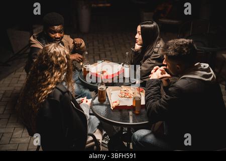 Eine Gruppe von Freunden genießt nachts Pizza und Getränke im Freien in gemütlicher Atmosphäre Stockfoto