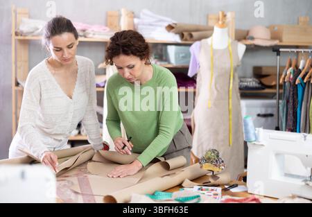 Zwei Schneiderinnen zeichnen eine Skizze der zukünftigen Kleidung auf Papier mit Mustern in der Nähwerkstatt Stockfoto