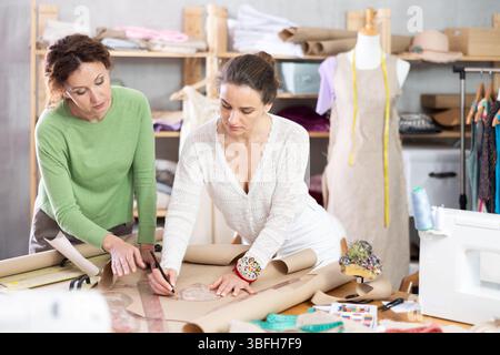 Zwei Schneiderinnen zeichnen eine Skizze der zukünftigen Kleidung auf Papier mit Mustern in der Nähwerkstatt Stockfoto