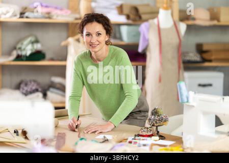 Die Schneiderin zeichnet in der Nähwerkstatt anhand von Mustern eine Skizze der zukünftigen Kleidung auf Papier Stockfoto
