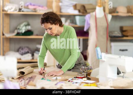 Die Schneiderin zeichnet in der Nähwerkstatt anhand von Mustern eine Skizze der zukünftigen Kleidung auf Papier Stockfoto