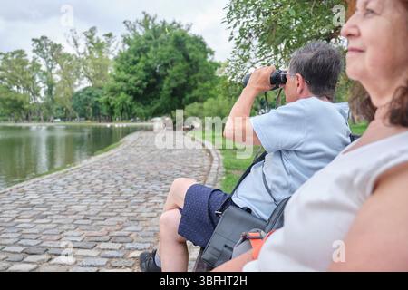Senior Hispanic-Paar, das sich in einem Park entspannt. Im Vordergrund blickt die Frau auf den See vor sich, hinter ihr benutzt der Mann ein Fernglas Stockfoto