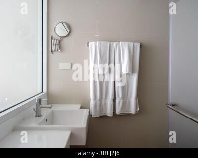Paare Set aus großen und kleinen sauberen weißen Handtüchern, gefaltet an einem Metallbügel an der Wand mit Fliesen in der Nähe von Keramikbecken und durchscheinendem Glasfenster i Stockfoto
