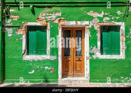 Eine lebhafte grüne Wand mit verwitterten Holzläden an zwei Fenstern und einer verzierten Vintage-Tür auf der Insel Burano, Italien. Stockfoto