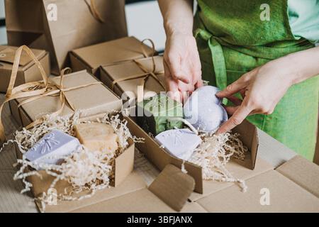 Die Person legt handgemachte Seifen und Lavendelzweige in eine Geschenkbox mit geschreddertem Füllstoff. Handwerkliche Hautpflege, umweltfreundliche Verpackung, handgefertigt Stockfoto