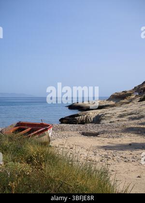 Redaktionelles Bild eines verwitterten Fischerbootes mit rot-weißer Färbung und teilweise griechischer Schrift an der Seite, die am Rand einer kleinen Bucht liegt. Stockfoto