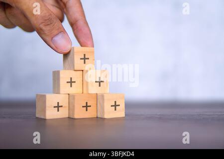 Eine Hand legt einen Holzwürfel mit einem schwarzen Pluszeichen auf eine Pyramide von Würfeln. Wachstum, positives, Wertschöpfungskonzept. Stockfoto
