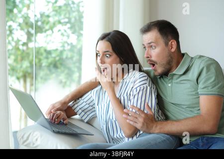 Ein überraschtes Paar, das zu Hause auf einer Couch sitzt und Inhalte eines PCs überprüft Stockfoto