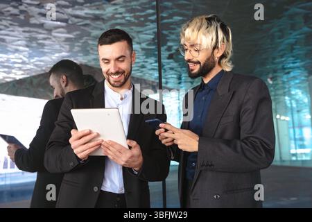 Zwei Geschäftsleute in Anzügen, die gemeinsam ein Tablet verwenden und im modernen Büro lächeln Stockfoto