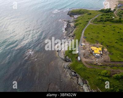 Aus der Vogelperspektive einer Baustelle an der Küste auf einer grünen Klippe am Meer. Zerklüftete Küste, Felsformationen und unvollendetes Haus mit gelbem Dach Stockfoto