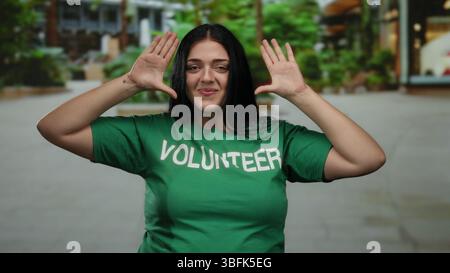 Frau in grünem Freiwilligenhemd posiert lächelnd in urbaner Outdoor-Umgebung mit Grün und modernen Gebäuden im Hintergrund, die Gemeinschaftsgeist und Gemeindegeist visualisieren Stockfoto