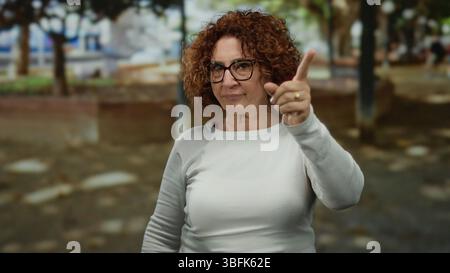 Frau gibt Missbilligung im Park, trägt Brille mit brünetten lockigen Haaren im Hintergrund im Freien, was auf einen nachdenklichen Moment während sunn hindeutet Stockfoto