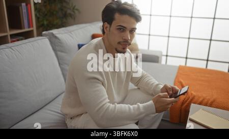 Hübscher junger hispanischer Mann, der das Telefon in einer gemütlichen Wohnzimmerwohnung benutzt, einen lässigen Pullover trägt, sieht zuversichtlich und entspannt drinnen aus, reflektiert modern Stockfoto