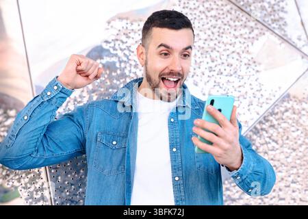 Aufgeregter junger Mann, der feiert, während er das Smartphone in heller Umgebung betrachtet Stockfoto