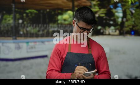 Junger hispanischer Mann in Brille macht Notizen in einer Parklandschaft, trägt ein rotes Hemd und eine Schürze, hält sich im Freien mit einem nachdenklichen Ausdruck fest, der aufwertet Stockfoto