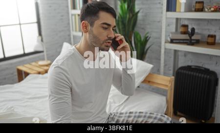 Junger hispanischer Mann, der auf dem Bett im Schlafzimmer sitzt und telefoniert in hellem, modernem Innenraum mit Koffer und Pflanzen im Hintergrund sichtbar ist und eine gemütliche schafft Stockfoto