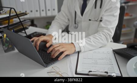 Junger Mann Arzt, der an Laptop in der Klinik Büro Innenraum arbeitet, konzentriert auf digitale Aufzeichnungen und Papierkram Management, schlägt Profess vor Stockfoto