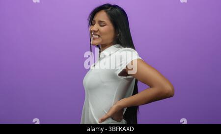 Die Frau mit Rückenschmerzen steht mit geschlossenen Augen vor einem leuchtend violetten Hintergrund, trägt ein weißes Hemd und vermittelt Beschwerden mit einem leichten Gramm Stockfoto