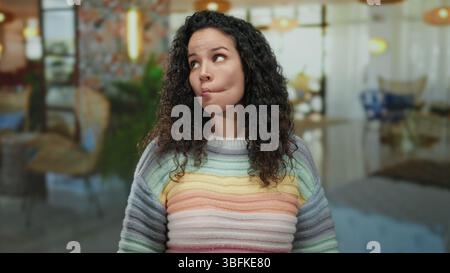 Hispanische Frau mit lockigem Haar, die drinnen in einem Hotel steht und einen farbenfrohen gestreiften Pullover trägt, der verschiedene nachdenkliche Posen gegen einen bl ausdrückt Stockfoto