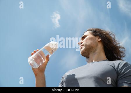 Hübsche lockige Brünette Mann tragen graues T-Shirt mit Wasserflasche vor blauem Himmel Hintergrund. Sommer Hitze Wetter. Im Freien. Hero-Ansicht. Nahaufnahme. Banne Stockfoto