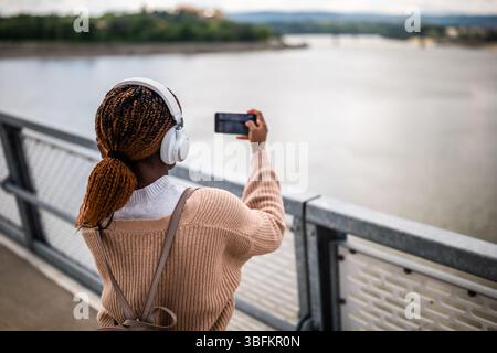 Glückliche junge schwarze Frau schaut in die Kamera und lächelt. Sie genießt einen sonnigen Tag im Freien und hört Musik, während sie Fotos macht. Stockfoto