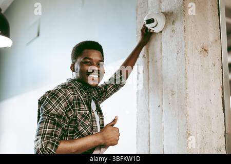 Mann, der eine Sicherheitskamera in einem modernen Bürobereich installiert, während er lächelt und die Daumen hochgibt Stockfoto