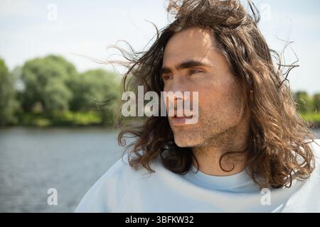 Porträt eines gut aussehenden jungen kaukasischen brünetten Mannes mit langen lockigen Haaren, der in der Nähe des Flusses steht. Sommerurlaub und Spaziergang. Langsam leben. Stockfoto
