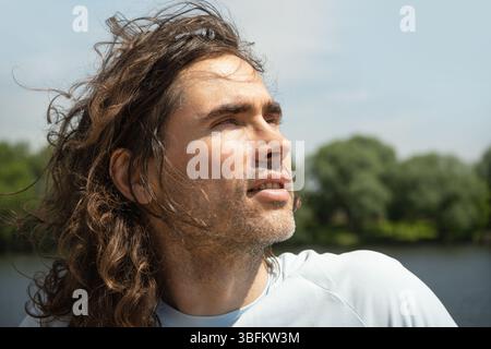 Porträt eines gut aussehenden jungen kaukasischen brünetten Mannes mit langen lockigen Haaren, der in der Nähe des Flusses steht, die Natur genießt und nach oben blickt. Sommerurlaub Stockfoto