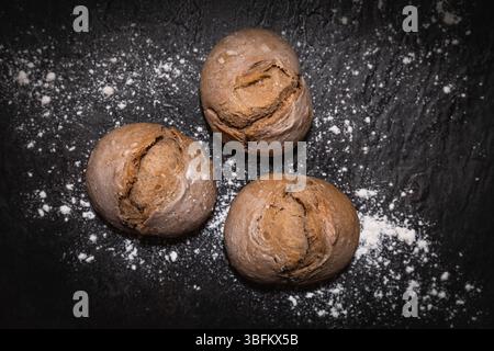Drei hausgemachte Sauerteigbrötchen mit rustikaler Kruste und Mehlstaubung, zentral auf dunkler Schieferoberfläche platziert, von oben in Studiobeleuchtung fotografiert. Stockfoto