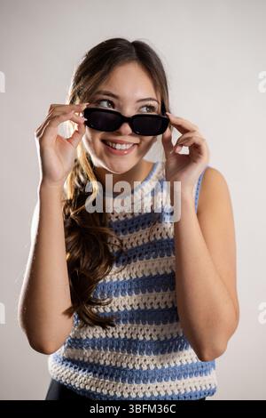 Studioaufnahme in Hüfthöhe einer jungen erwachsenen Frau, die über dem Rahmen lächelt, während sie die Kanten ihrer Sonnenbrille hält. Sie hat lange brünette Haare und trägt einen Stockfoto