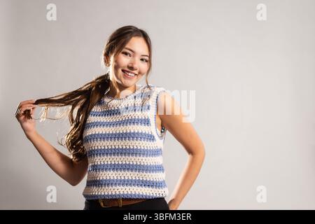Studio-Aufnahme in der Taille von einer jungen erwachsenen Frau, die direkt in die Kamera lächelt, mit einer Hand auf der Hüfte und der anderen das Ende ihres Pferdeschwanzes hält Stockfoto