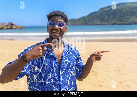 Junger Mann, der mit Zeigefingern auf sich und zur Seite zeigt, während er an einem sonnigen Strand lächelt Stockfoto
