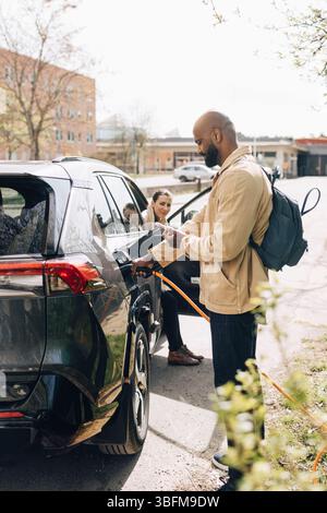 Frau, die auf einen Mann blickt, der ein Elektroauto auflädt, während er auf der Straße ein Smartphone benutzt Stockfoto