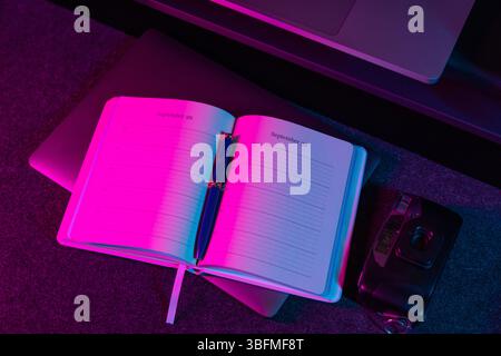 Ein offener Planer oder Tagebuch, in dem der „29. September“ und der „September 30“ mit einem Stift dargestellt werden, beleuchtet durch leuchtende rosa und blau/violette Neonlichter. Ein Laptop und Stockfoto