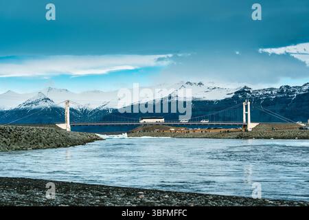 Malerischer Blick auf die berühmte Hängebrücke mit einer Autofahrt über die Gletscherlagune Jokulsarlon und Bergkette im Hintergrund im Winter bei Southeast Icel Stockfoto